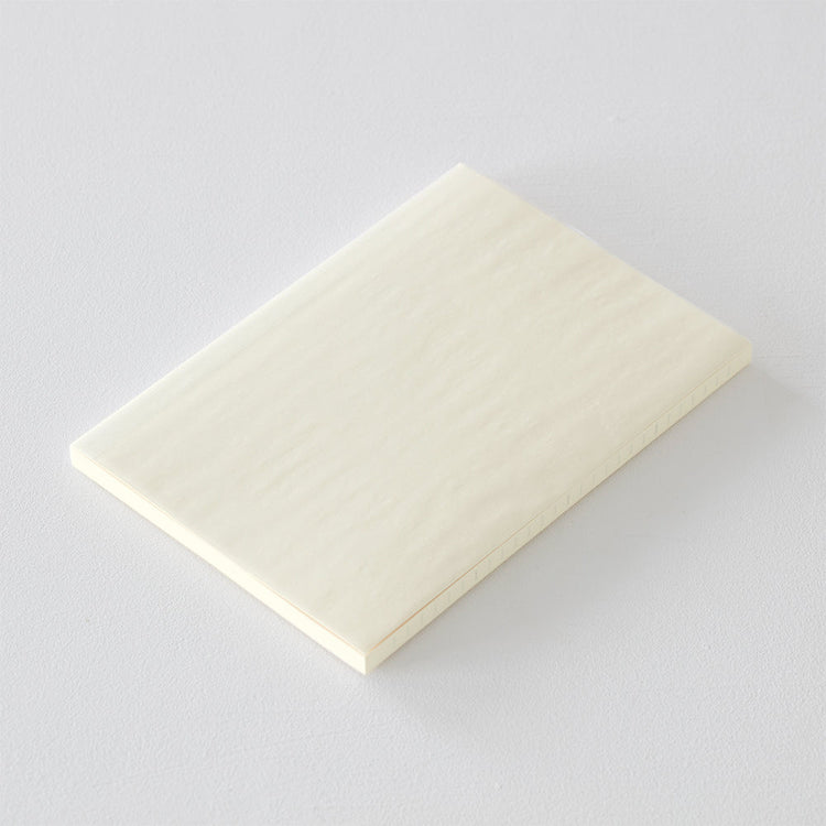 A5 MD Notebook Diary 2026 with a minimalist cream-toned glassine paper cover, thread-stitched binding, and a white bookmark string. The open spread displays a block-style monthly calendar on one side and lined memo pages on the other, printed on smooth MD PAPER designed for a premium writing experience.