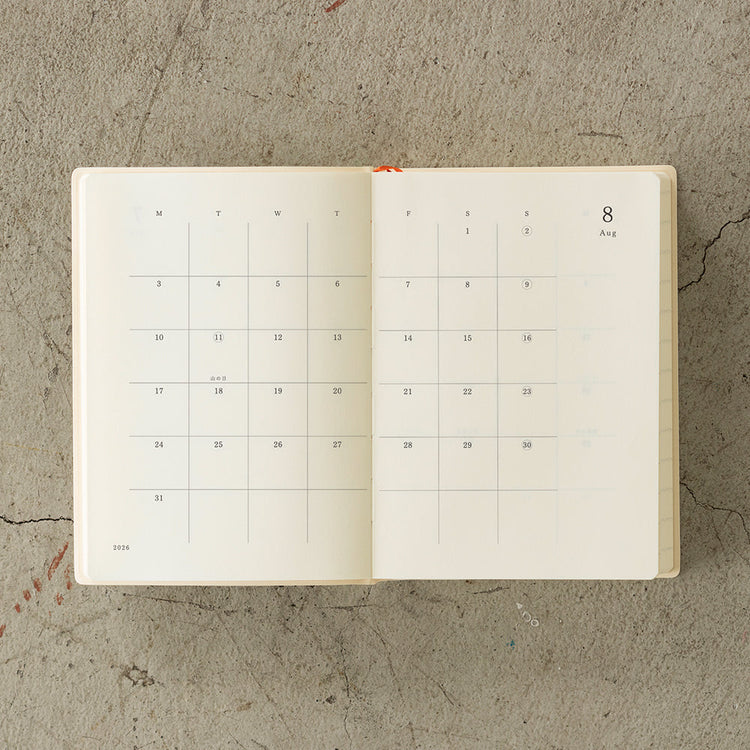 MD Notebook Diary (A6) 1 Day 1 Page 2026 with a minimalist PVC cover, featuring a year stamp on the spine, two bookmark strings, and monthly index tabs. The diary is open to reveal its premium MD PAPER with a clean, spacious layout for daily entries.