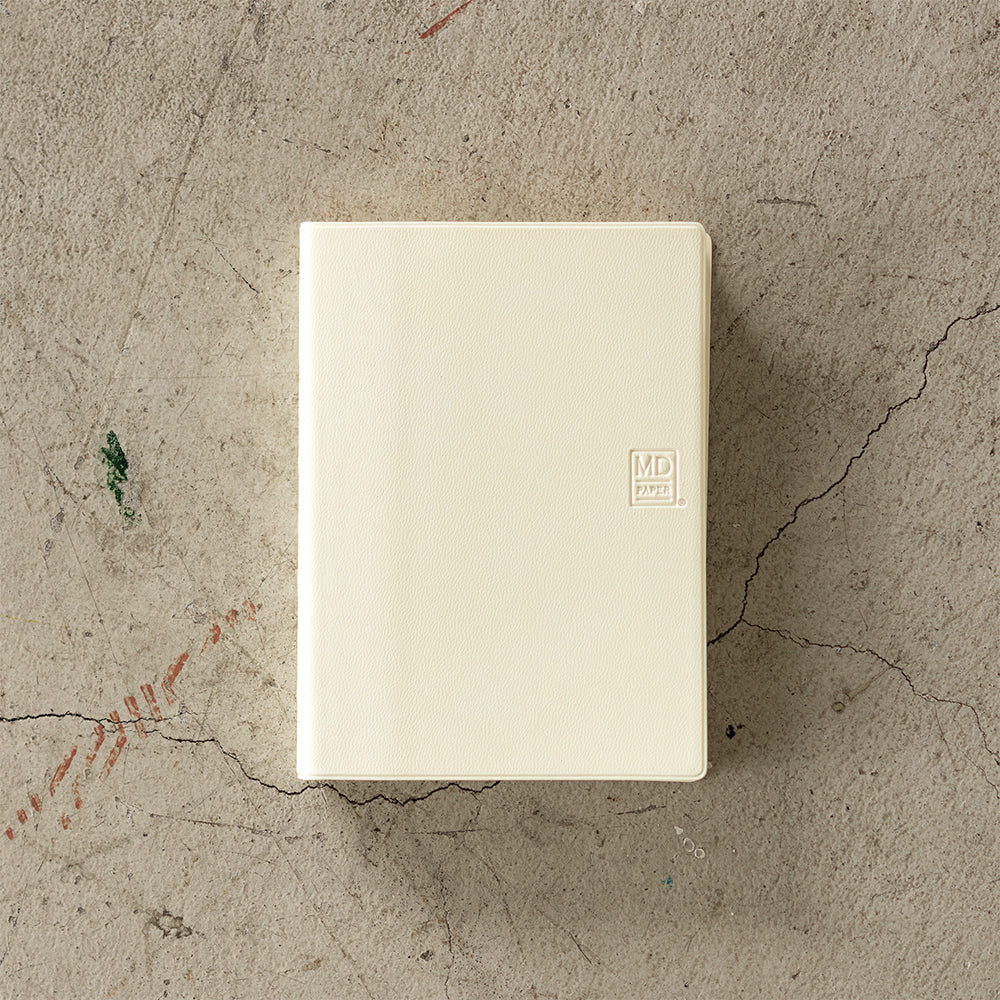 MD Notebook Diary (A6) 1 Day 1 Page 2026 with a minimalist PVC cover, featuring a year stamp on the spine, two bookmark strings, and monthly index tabs. The diary is open to reveal its premium MD PAPER with a clean, spacious layout for daily entries.