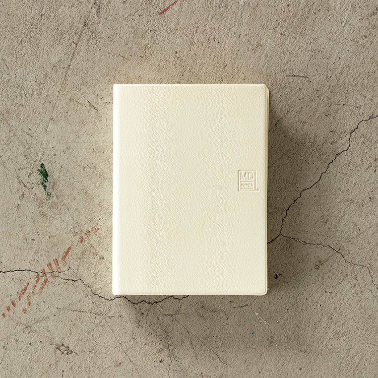 MD Notebook Diary (A6) 1 Day 1 Page 2026 with a minimalist PVC cover, featuring a year stamp on the spine, two bookmark strings, and monthly index tabs. The diary is open to reveal its premium MD PAPER with a clean, spacious layout for daily entries.