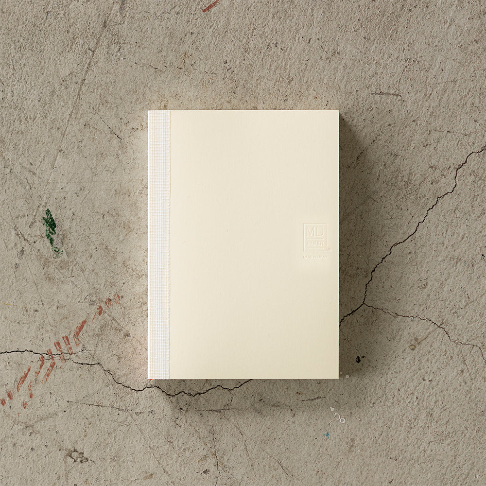 A6 MD Notebook Diary 2026 with a minimalist cream glassine paper cover, featuring monthly block schedule pages, weekly planning sections divided into eight segments, and blank memo pages. Thread-bound for 180-degree flat opening, complete with a slim bookmark string and index label.