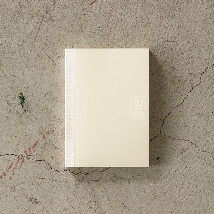 A6 MD Notebook Diary 2026 with a minimalist cream glassine paper cover, featuring monthly block schedule pages, weekly planning sections divided into eight segments, and blank memo pages. Thread-bound for 180-degree flat opening, complete with a slim bookmark string and index label.