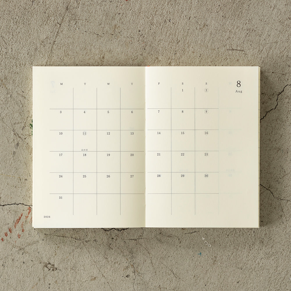 A6 MD Notebook Diary 2026 with a minimalist cream glassine paper cover, featuring monthly block schedule pages, weekly planning sections divided into eight segments, and blank memo pages. Thread-bound for 180-degree flat opening, complete with a slim bookmark string and index label.