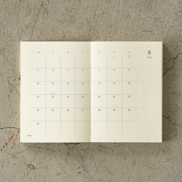 A6 MD Notebook Diary 2026 with a minimalist cream glassine paper cover, featuring monthly block schedule pages, weekly planning sections divided into eight segments, and blank memo pages. Thread-bound for 180-degree flat opening, complete with a slim bookmark string and index label.