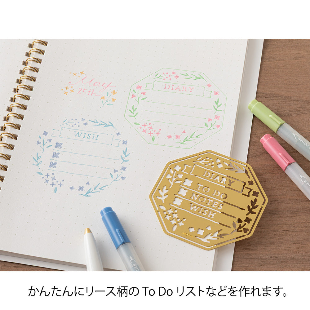 A brass stencil with floral and leafy cut-outs forming wreath-style designs, along with headings, to-do boxes, and animal details for decorating planners and memos.