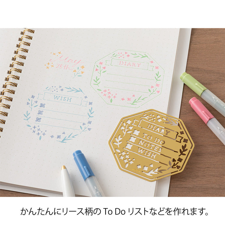 A brass stencil with floral and leafy cut-outs forming wreath-style designs, along with headings, to-do boxes, and animal details for decorating planners and memos.