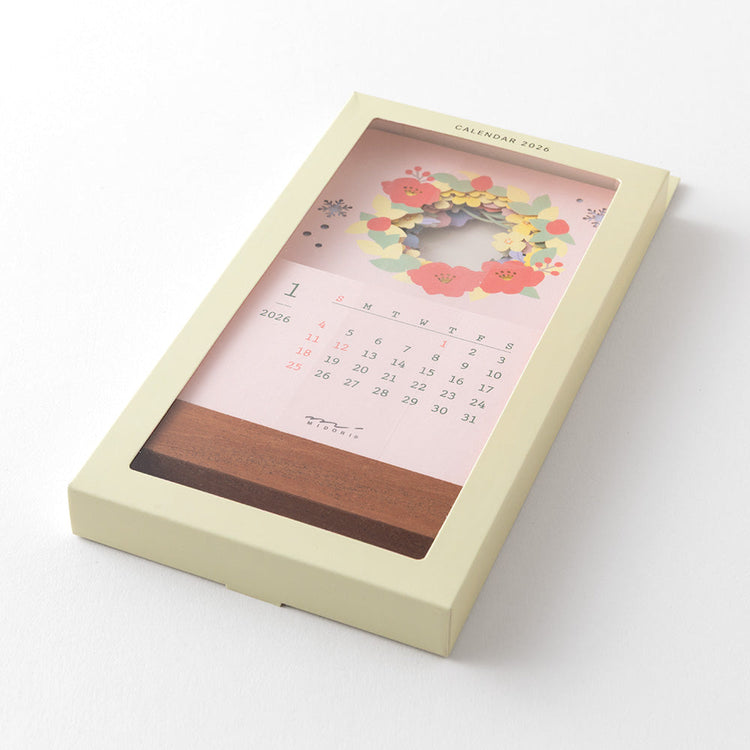 A delicate 2026 desktop calendar featuring layered, laser-cut floral designs that change with the seasons. Each month reveals a new arrangement of blossoms, displayed on a compact wooden stand, bringing an artful and elegant touch to any desk or small space.