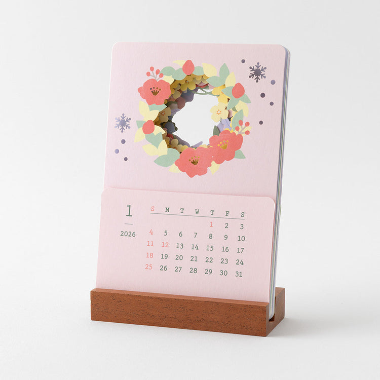 A delicate 2026 desktop calendar featuring layered, laser-cut floral designs that change with the seasons. Each month reveals a new arrangement of blossoms, displayed on a compact wooden stand, bringing an artful and elegant touch to any desk or small space.