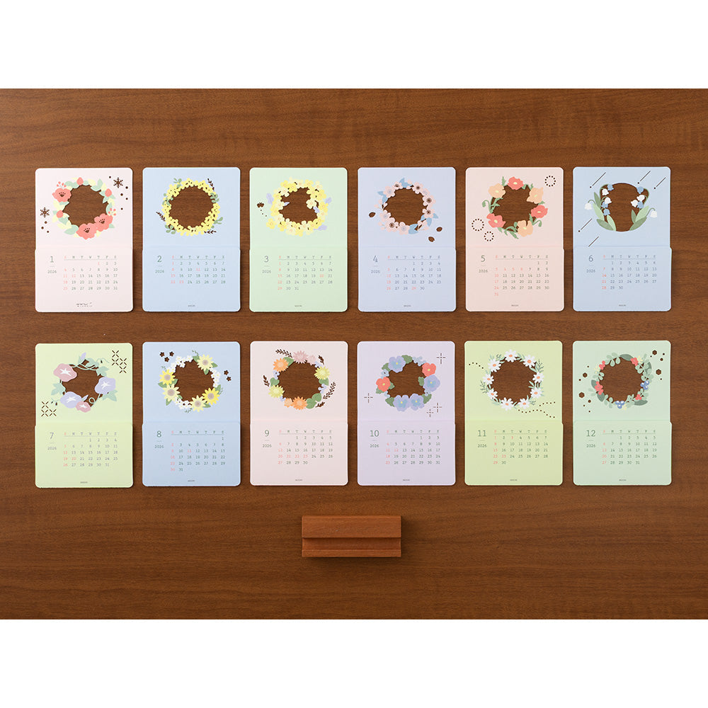A delicate 2026 desktop calendar featuring layered, laser-cut floral designs that change with the seasons. Each month reveals a new arrangement of blossoms, displayed on a compact wooden stand, bringing an artful and elegant touch to any desk or small space.