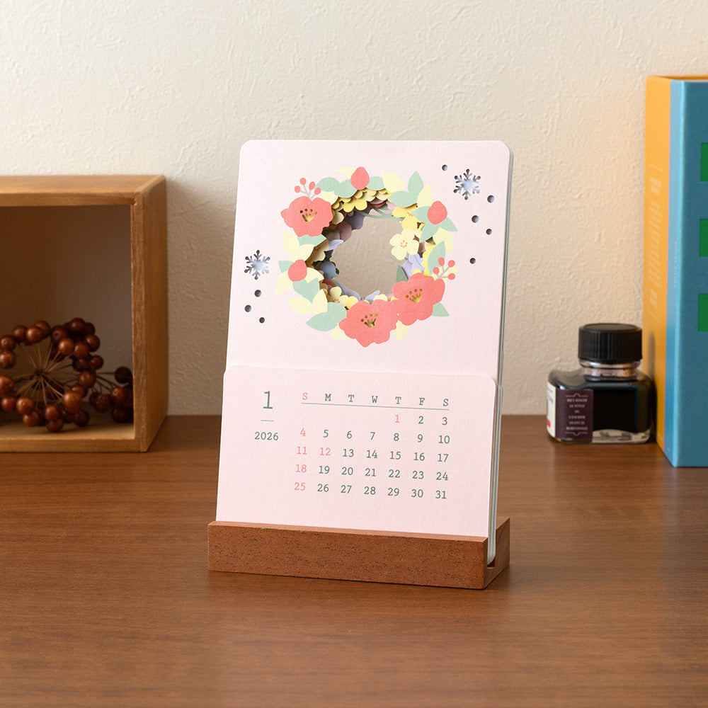 A delicate 2026 desktop calendar featuring layered, laser-cut floral designs that change with the seasons. Each month reveals a new arrangement of blossoms, displayed on a compact wooden stand, bringing an artful and elegant touch to any desk or small space.